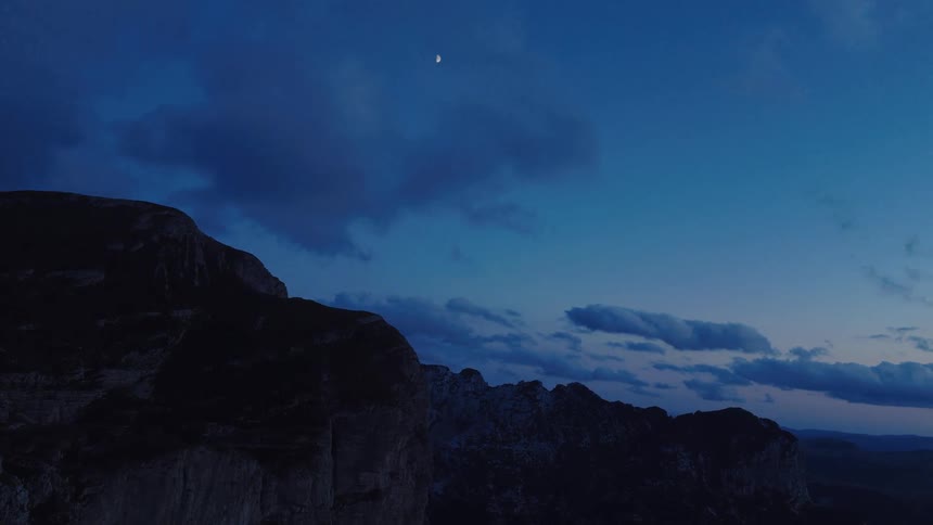 A sliver of the moon shines in the deep blue twilight sky above a dark, silhouetted mountain range