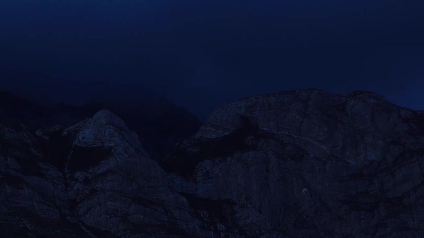 Static shot of a moody, dark mountain range under the deep blue night sky as fog rolls in