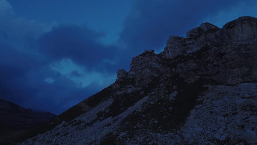 A slow aerial approach towards a rugged mountain peak under a deep blue, cloudy sky at dusk