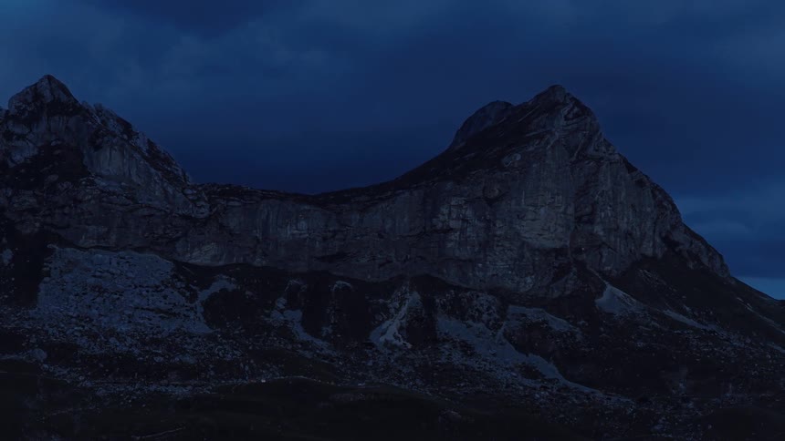 An epic aerial shot slowly pulls back from a dramatic mountain range under a dark blue, cloudy night sky