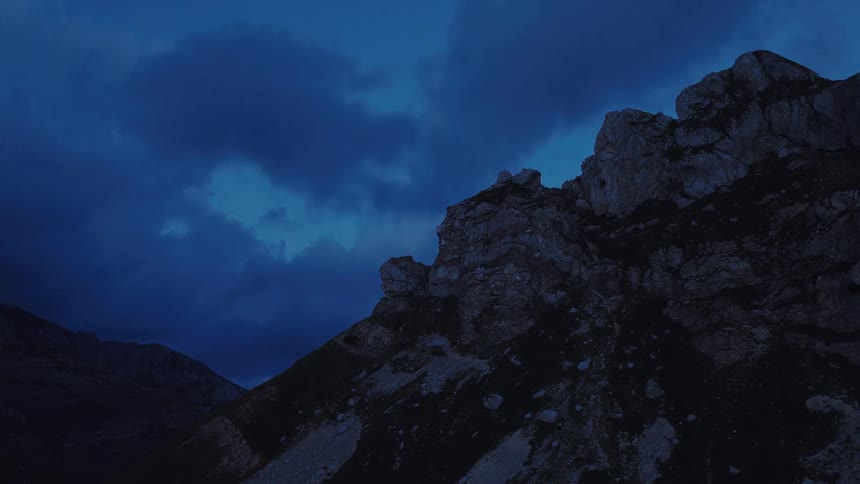 A slow aerial pullback reveals a rugged mountain peak under a dark and moody blue sky at dusk