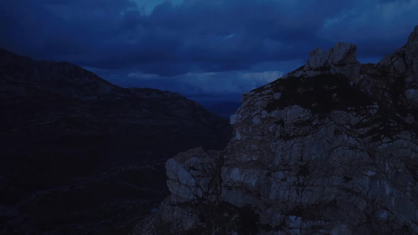 An ominous aerial shot slowly pans over a rugged mountain peak to reveal a dark valley at twilight