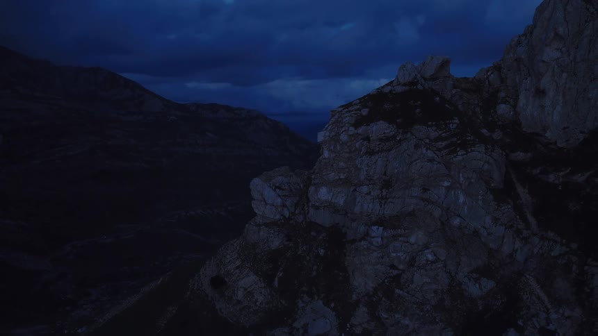 A slow aerial shot reveals a vast and rugged mountain landscape under a dark, cloudy sky at twilight
