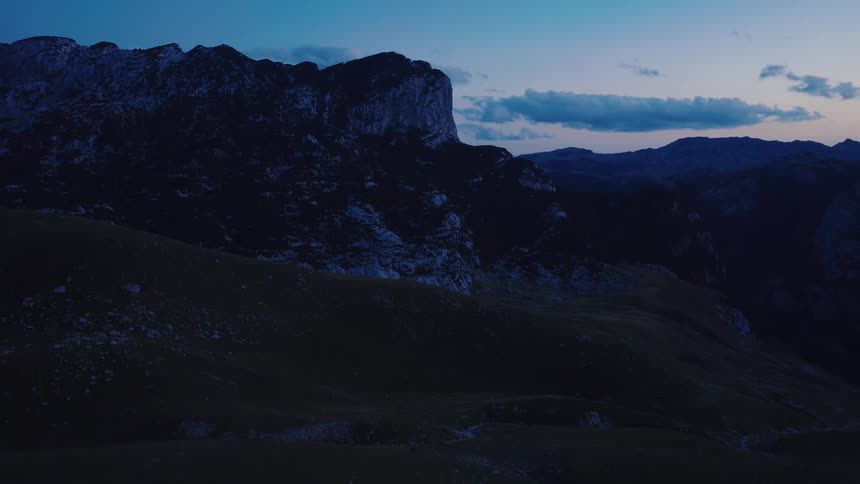 Aerial view flying backward over a dark, grassy mountain landscape with majestic peaks against a deep blue twilight sky