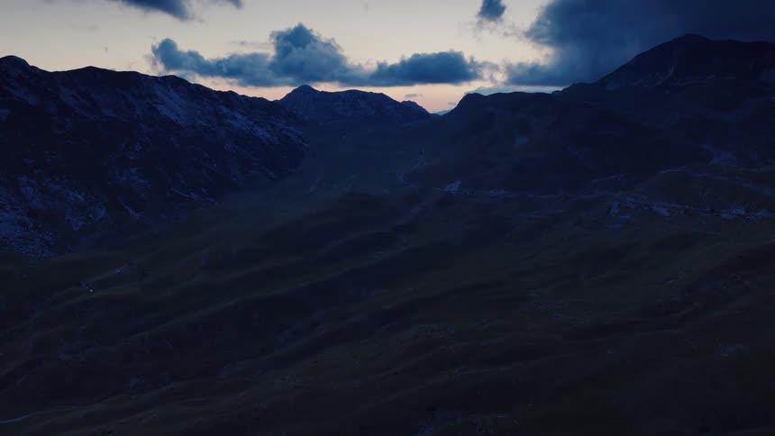 A drone slowly flies over a dark, rolling mountain landscape during the blue hour just after sunset