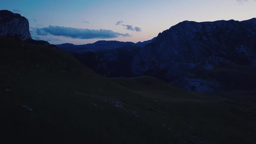 An aerial view slowly rises over a dark, grassy valley at dusk to reveal a majestic mountain range against the twilight sky