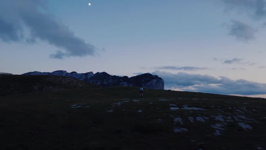 A lone runner traverses a vast mountain landscape at dusk under the light of the moon