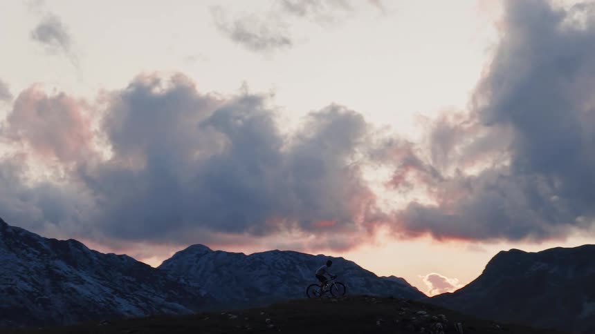The silhouette of a mountain biker rides along a high-altitude ridge with a backdrop of snowy mountains and a dramatic, cloudy sunset