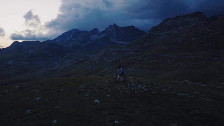 An aerial shot follows a mountain biker riding across a vast, grassy mountain range under a dark, cloudy sky