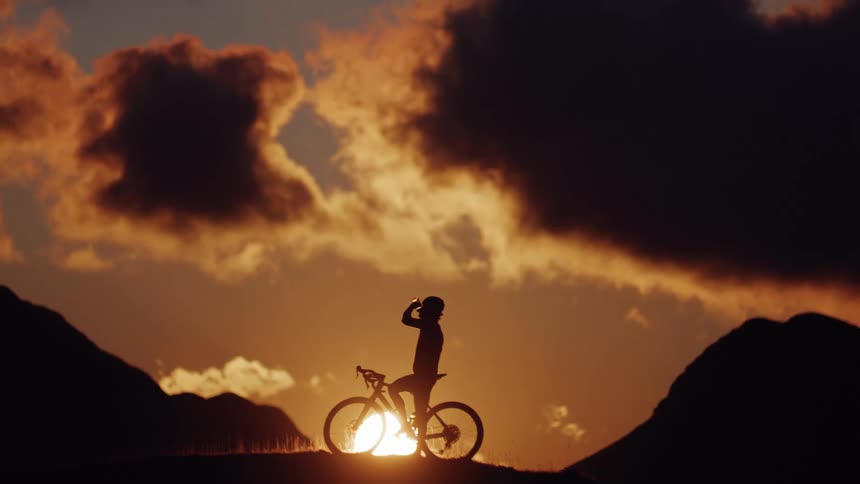 A silhouetted cyclist takes a water break on a mountain ridge during a dramatic golden sunset