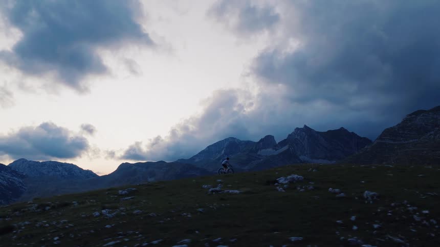 A lone mountain biker pauses on a grassy ridge, overlooking a vast mountain range under a dramatic and moody sky at dusk