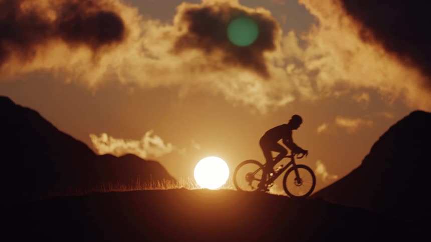 A cyclist rides on a mountain ridge silhouetted against a dramatic golden sunset