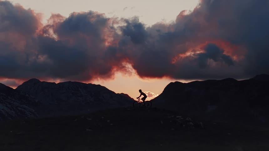 A mountain biker rides slowly across a mountain ridge, silhouetted against a dramatic and fiery sunset