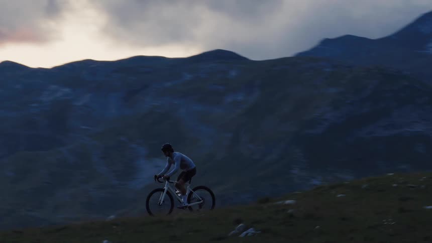 A lone cyclist pedals uphill on a grassy mountain pass during the blue hour at dusk