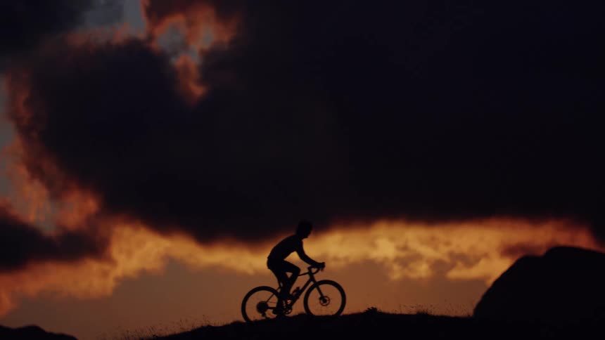 A silhouetted cyclist rides in slow motion along a mountain ridge against a dramatic and fiery sunset sky