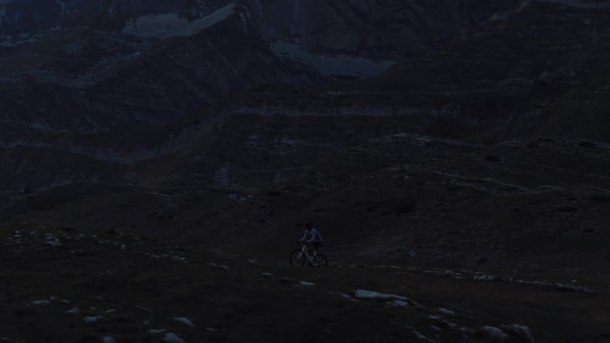 A lone mountain biker is silhouetted against a vast, dark mountain range while riding up a grassy hill under a moody sky