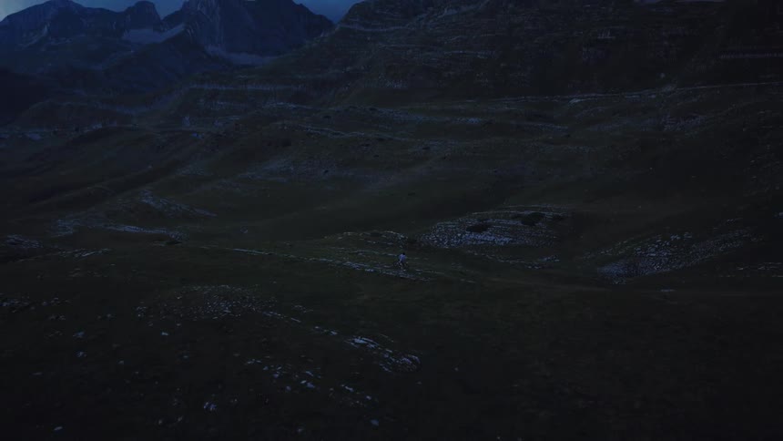 A dramatic aerial view follows a lone hiker traversing a vast and moody mountain landscape at twilight