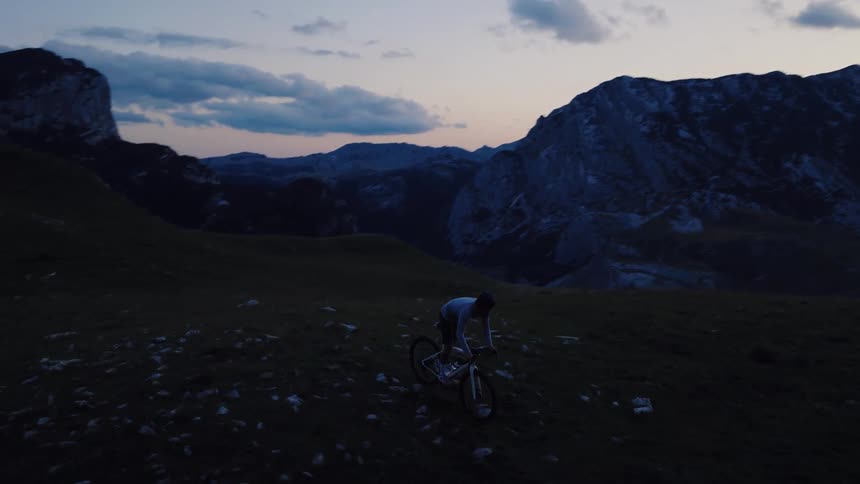 An aerial view follows a silhouetted mountain biker riding along a scenic alpine ridge at dusk