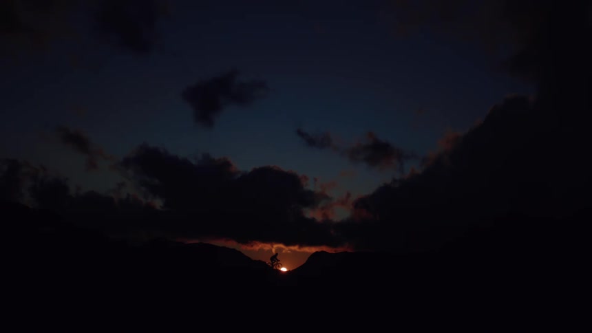 A silhouetted mountain biker slowly rides along a mountain ridge in front of a dramatic sunset