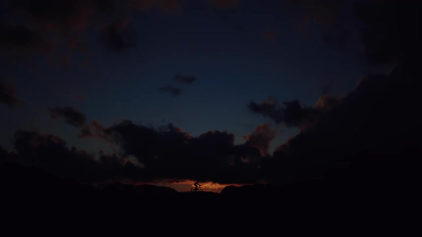 A lone cyclist rides along a silhouetted mountain ridge against a dramatic sunset sky