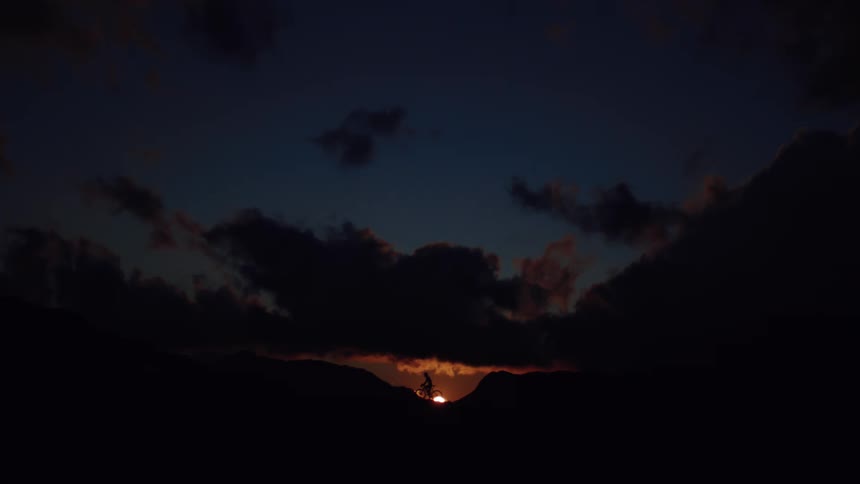 Dramatic silhouette of a mountain biker riding on a mountain ridge against the setting sun and dark clouds