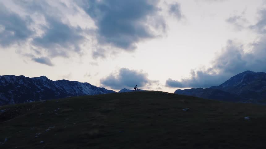 Epic aerial shot follows a mountain biker as they ascend a grassy peak against a dramatic mountain landscape at dusk