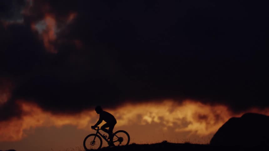 A cyclist rides in slow motion across a hilltop, silhouetted against a dramatic and fiery sunset sky
