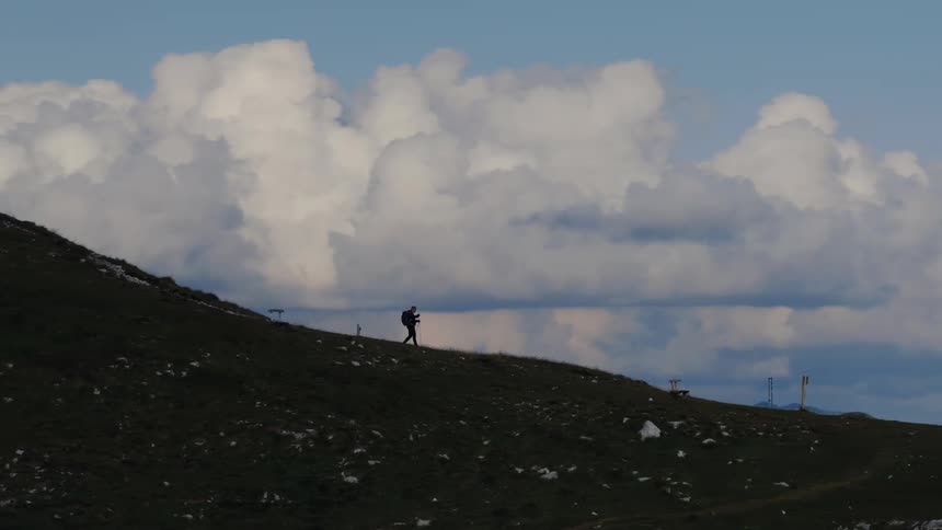 A lone hiker ascends a grassy mountainside, silhouetted against a vast and cloudy sky