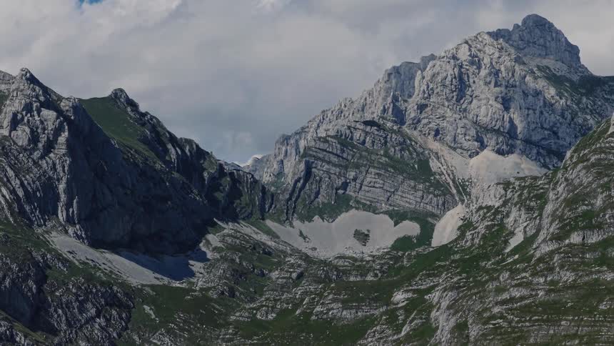 A slow zoom out reveals a vast and rugged alpine mountain range with shadows from clouds drifting across the peaks