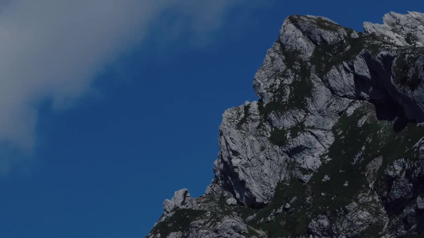 A majestic rocky mountain peak is revealed in a slow vertical tilt against a vibrant blue sky with passing clouds