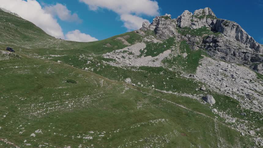 An aerial drone shot captures a lone vehicle driving up a steep, green mountain slope towards a rocky summit on a sunny day