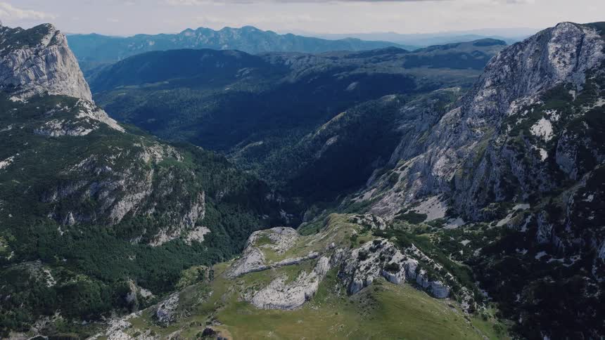 A stunning aerial view pulls back from a deep mountain valley revealing the vast, rocky, and forested landscape