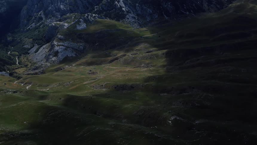 A static aerial shot captures the dramatic play of sunlight and shadow across rolling green hills at the base of a rugged mountain range