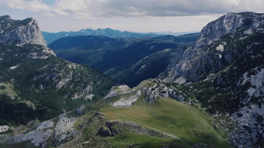 Slow aerial flight over a vast, green mountain valley surrounded by rugged, rocky peaks under a partly cloudy sky
