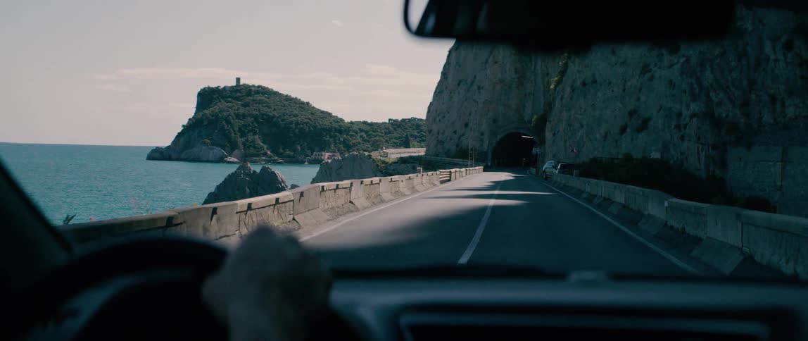 POV of driving a car along a winding and scenic coastal road next to the ocean before entering a tunnel through a mountain