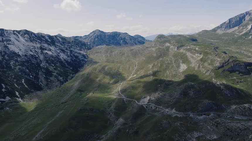 A static aerial shot captures the vast and rugged landscape of a green mountain range with a winding road under a bright, clear sky