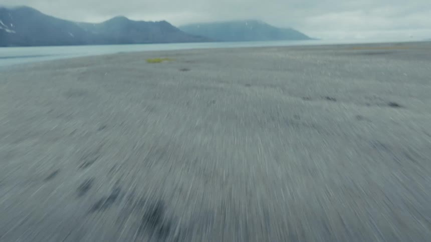 Low aerial shot speeds over a vast, barren landscape, tilting up to reveal misty mountains and a fjord on an overcast day