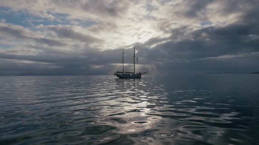 A majestic sailboat floats peacefully on a calm ocean as heavenly sun rays break through a dramatic cloudy sky
