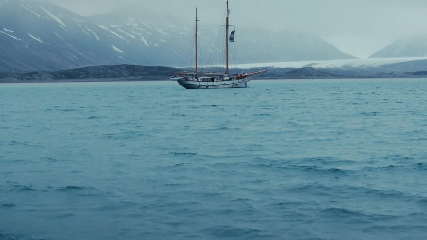 A slow tilt up from the cold blue ocean reveals a classic schooner anchored in a remote arctic bay with glaciers and snowy mountains in the background