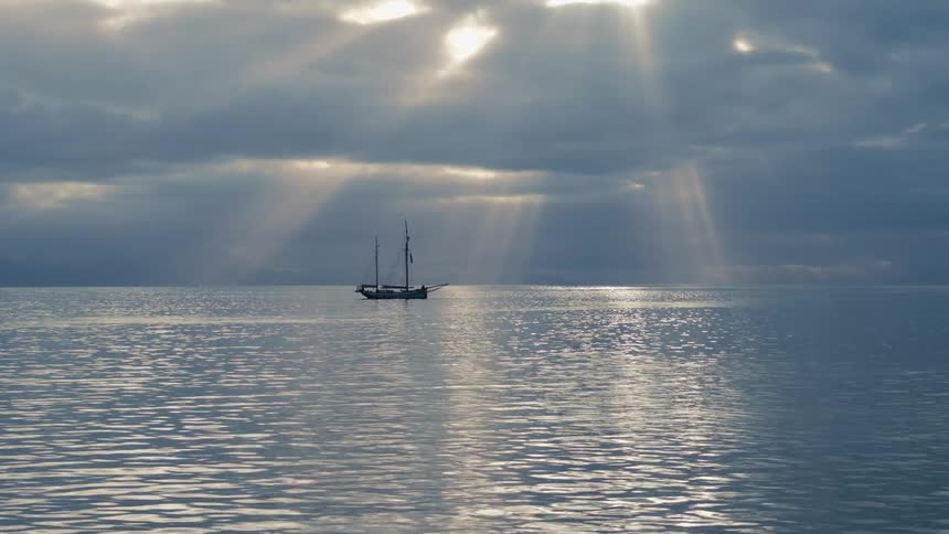 A lone sailboat floats on a calm ocean as heavenly sun rays break through the clouds