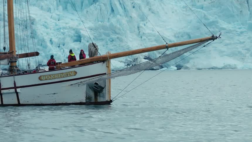 A classic wooden sailboat with a crew aboard floats peacefully in the arctic waters before a towering wall of glacial ice