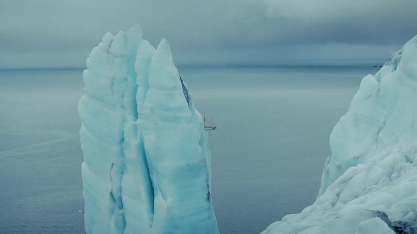 A solitary sailboat is framed between two enormous blue icebergs as it glides across the calm arctic ocean