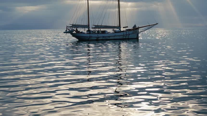 A serene view of a classic sailboat anchored on the calm ocean, with dramatic sunbeams piercing through the clouds