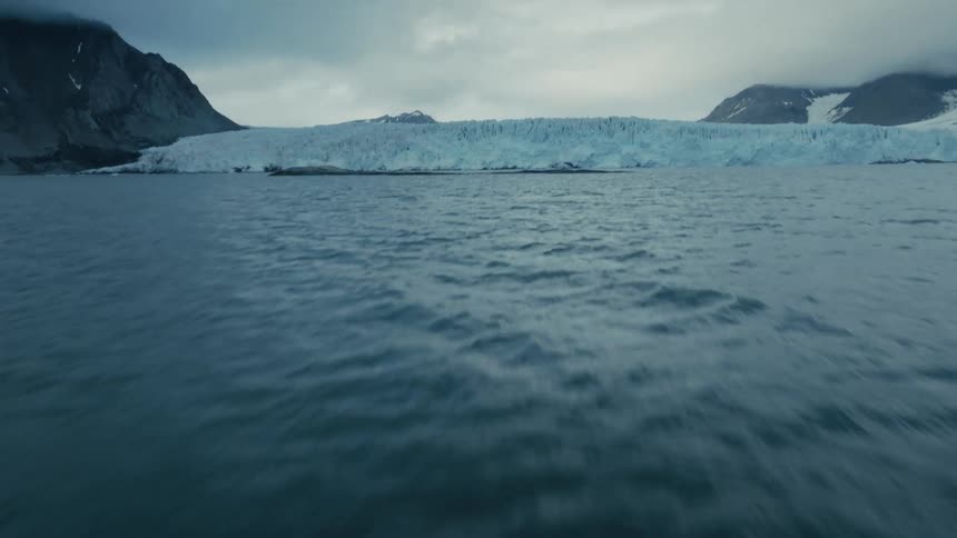 A dramatic aerial shot flies low and fast over the arctic ocean, revealing a massive glacier between rugged mountains