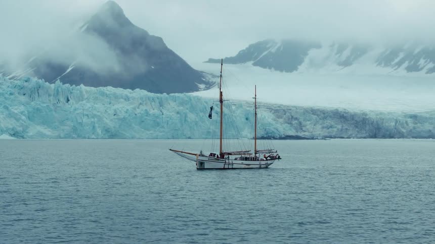 A majestic sailboat drifts through a tranquil polar bay with a towering blue glacier and fog-enshrouded mountains in the background