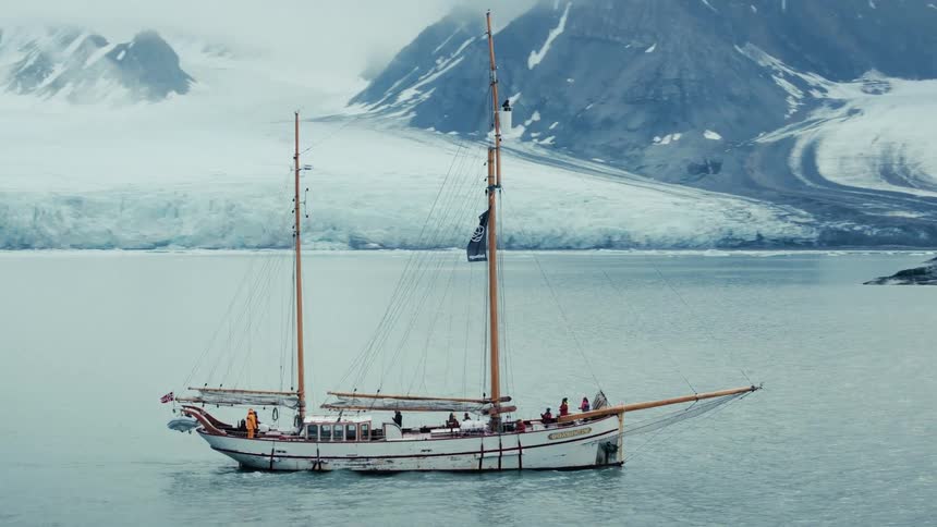 An aerial view shows a classic schooner sailboat navigating through an icy bay with a massive glacier and snowy mountains in the background