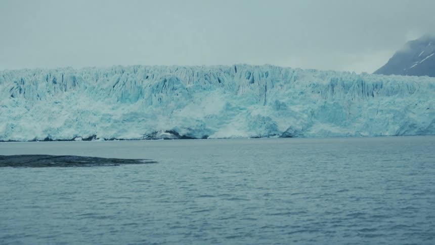 A slow tilt up from the cold Arctic ocean reveals a massive glacier on an overcast day