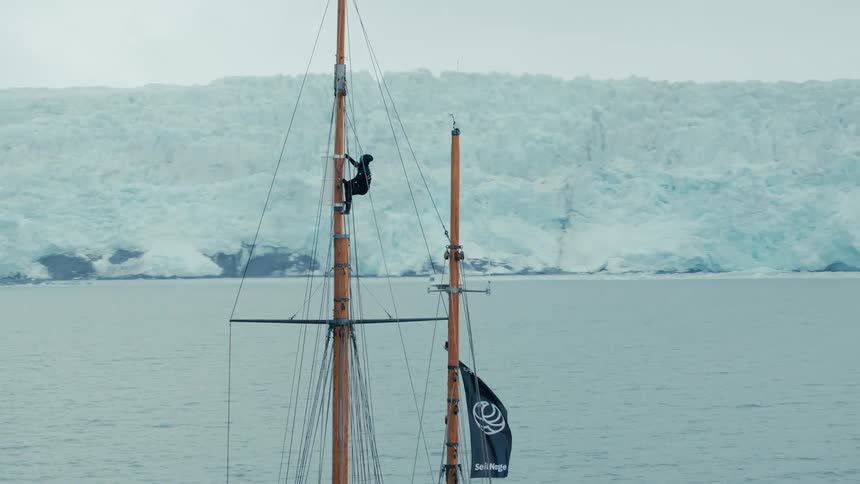 A sailor bravely climbs the tall wooden mast of a ship sailing through a frigid arctic bay with a massive glacier in the background