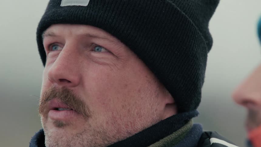 A close-up of a rugged man with a mustache looking around with a pensive expression on a cold, overcast day