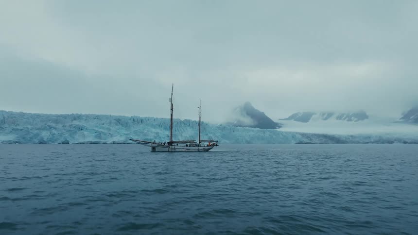 A beautiful sailboat cruises through the cold arctic sea with a vast glacier and snowy mountains in the background